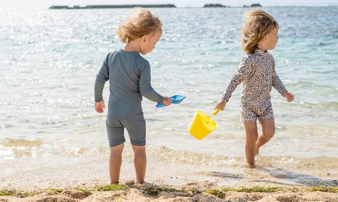duas crianças a brincar com um balde amarelo na praia vestidas com roupa que protege do sol