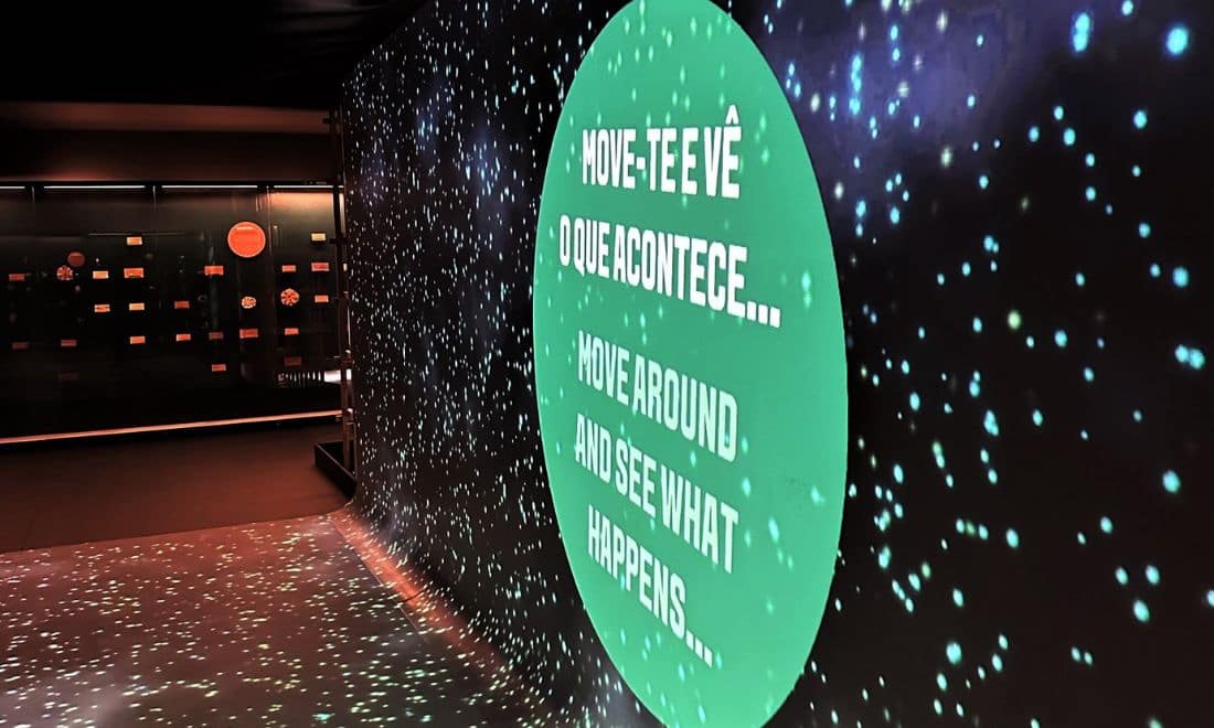 sala escura com um ecrã verde que convida os participantes a interagir