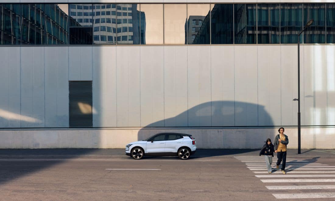 Automóvel Volvo EX30 à frente de um edifício com uma grande sombra projetada na parede e uma pai e uma filha de mãos dadas a atravessar a estrada