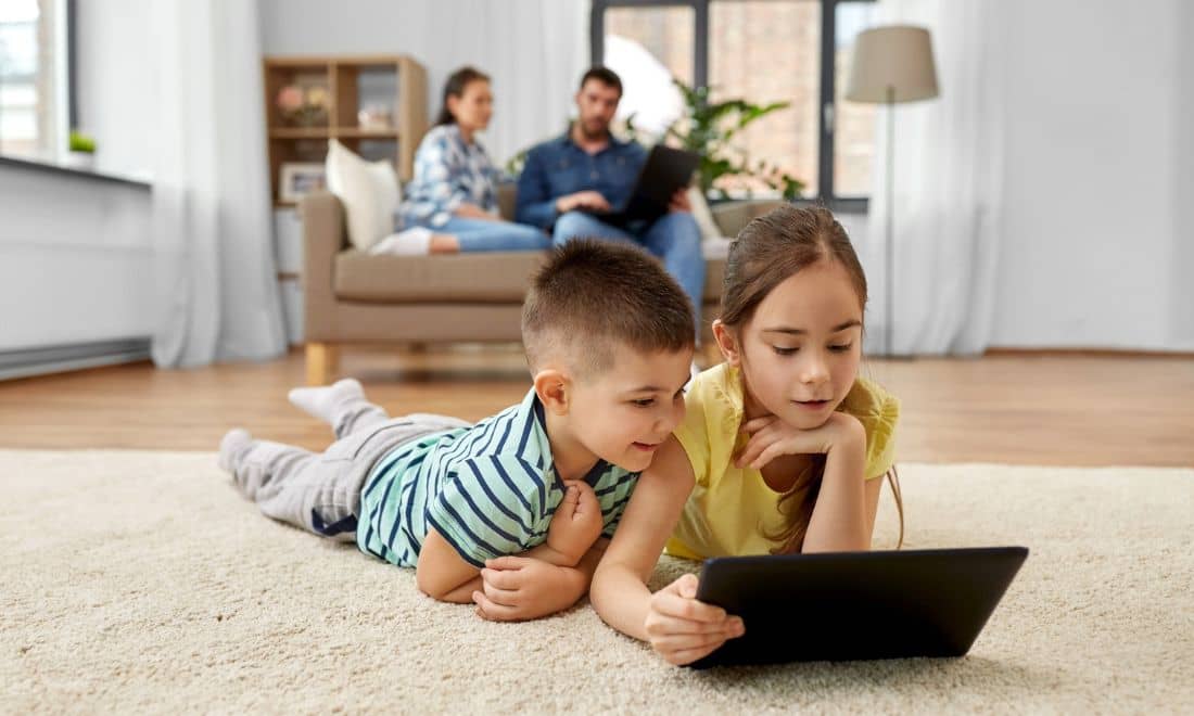 Competências digitais menina e menino no tablet no tapete da sala com pais ao fundo com computador na mão