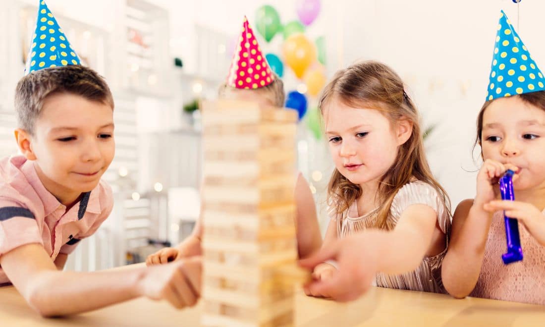 Festas de Aniversário crianças a jogar Jenga dentro de casa com chapéus de festa e balões ao fundo
